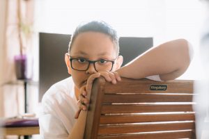 boy in white shirt wearing black framed eyeglasses. He is sitting with his face towards the back of the chair and looks lonely or sad.