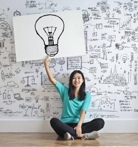A woman of Asian ethnicity is sitting on the floor holding up an image of a lightbulb. On the wall behind her are different images representing ideas, plans, and ways of organizing information. Music therapy is one way to work with executive functioning.