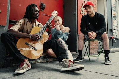 An image of three people seated on the sidewalk. A Black male-appearing person is playing the guitar while a white female-appearing person and a white-male appearing person sit beside him.